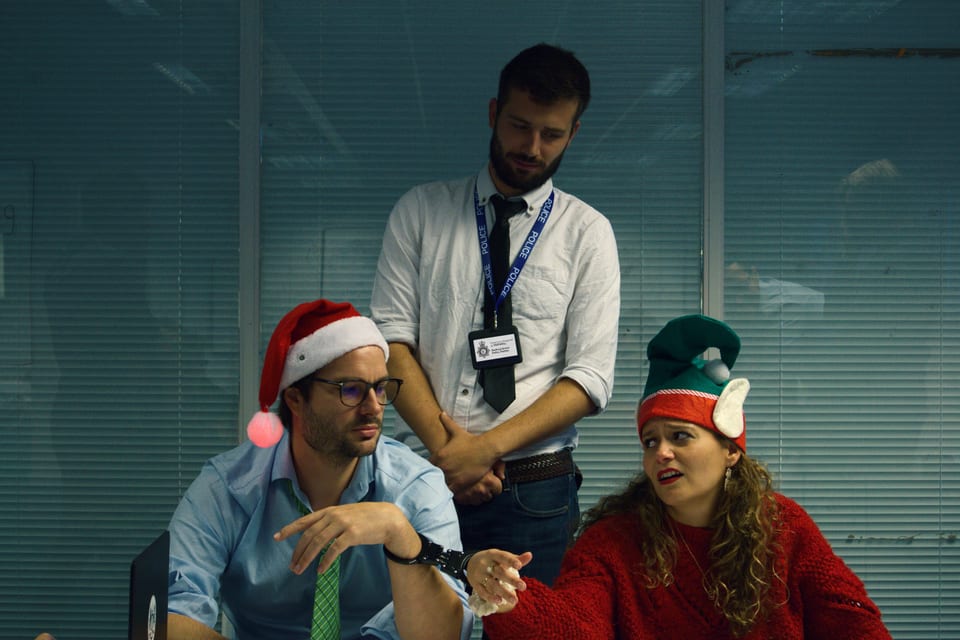 A detective questions two witnesses wearing Christmas hats in Death at the Office Party.